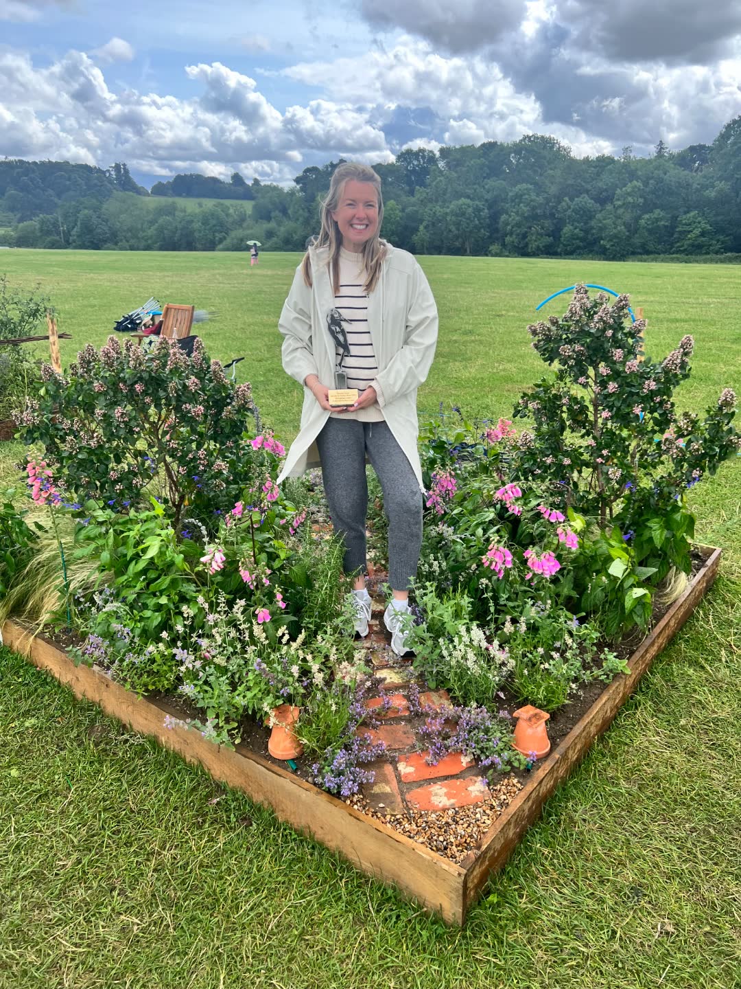 Emma Atkinson holding her award inside her completed show garden at Belvoir Castle Garden & Flower Show, with pink foxgloves and naturalistic planting around a brick path.
